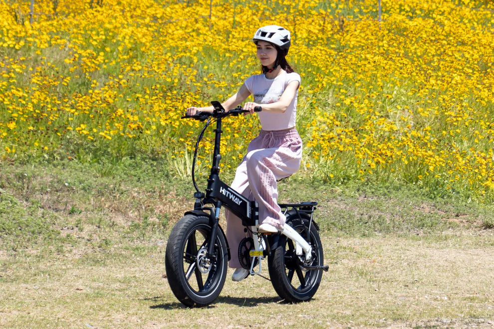Elektrische vouwfiets HITWAY BK10S, zwart wit, zijaanzicht schuin rechtsvoor, vrouw met fietshelm op op de fiets, veld met gele bloemen op de achtergrond